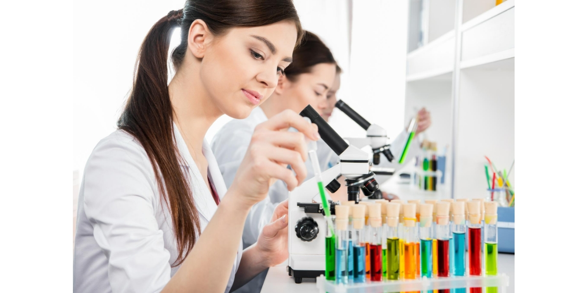 Due giovani scienziate al lavoro in laboratorio. Una di loro osserva attentamente al microscopio, mentre l'altra misura liquidi colorati in provette, evidenziando un ambiente di ricerca scientifica.