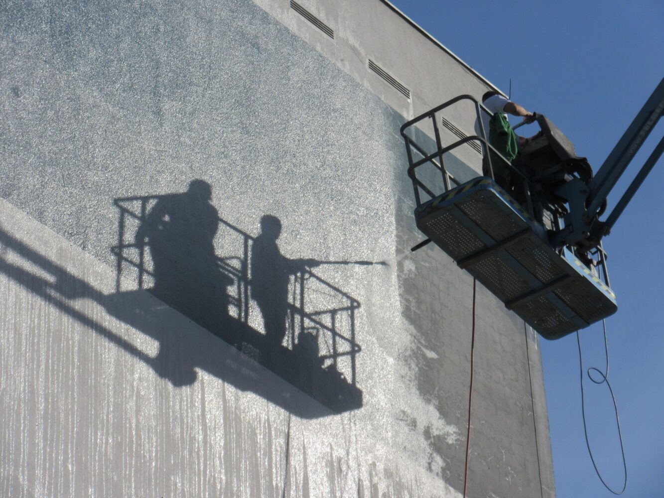 Due operai su una piattaforma elevatrice puliscono un muro, mentre le loro ombre si proiettano sul lato del palazzo in un giorno di sole.