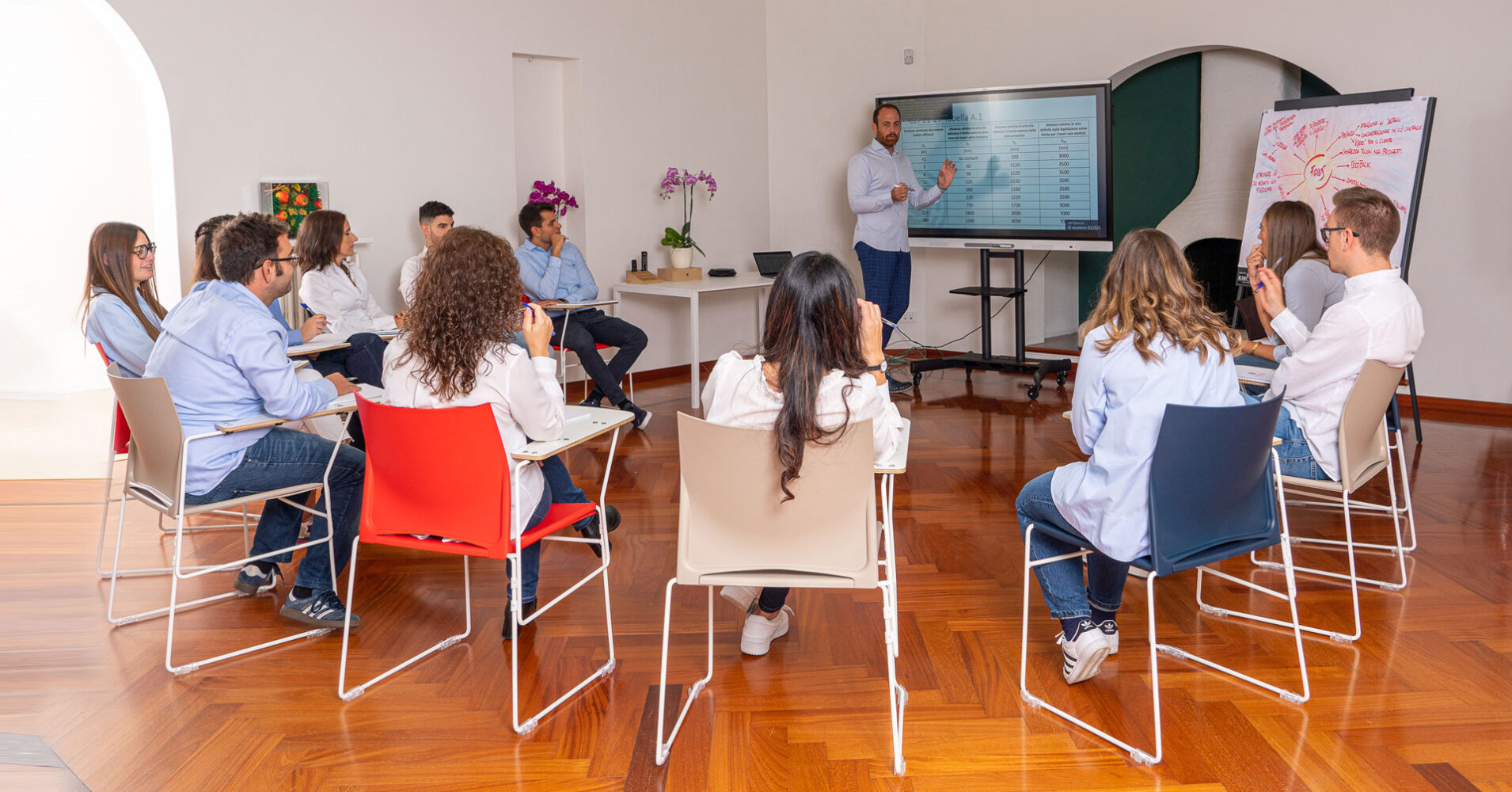 Un gruppo di professionisti in una sala riunioni ascolta un relatore che presenta dati su uno schermo. L'ambiente è moderno e luminoso.