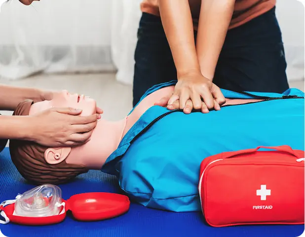Un manichino utilizzato per la pratica della rianimazione cardiopolmonare, mentre una persona esegue le compressioni toraciche. Accanto, una valigetta di pronto soccorso e attrezzature per la rianimazione.