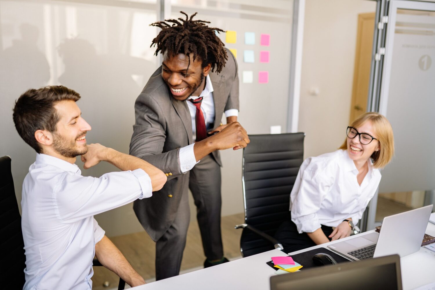 Un incontro di lavoro in ufficio, con tre persone sorridenti. Due uomini si stanno stringendo il gomito in segno di saluto, mentre una donna osserva divertita, circondati da attrezzature da ufficio.