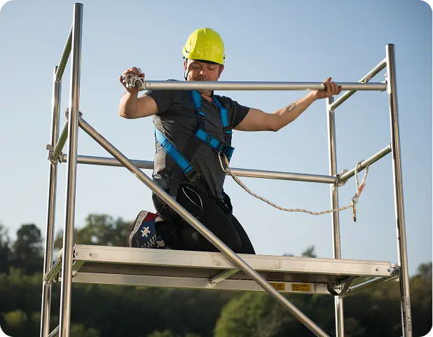 Un lavoratore su un'impalcatura sta installando un componente metallico. Indossa un caschetto giallo e una imbracatura di sicurezza, evidenziando l'importanza della sicurezza nei lavori in quota.