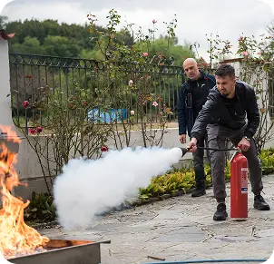 Un uomo utilizza un estintore per spegnere un incendio controllato, mentre un altro uomo osserva. Siamo in un ambiente esterno con piante e fiori.
