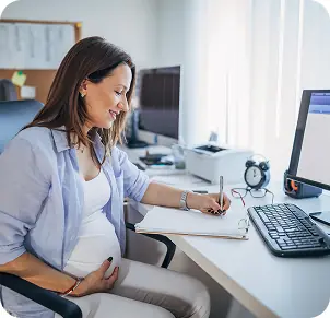 Donna incinta seduta alla scrivania, mentre scrive su un quaderno e guarda il computer. Atmosfera di lavoro serena.