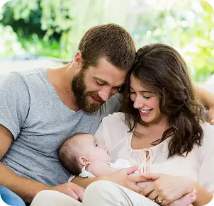 Una giovane coppia sorridente tiene in braccio il loro neonato, immersi in un'atmosfera serena e naturale. Il genitore maschile ha una folta barba e la madre indossa una camicia chiara. Sullo sfondo si intravedono piante verdi, creando un ambiente intimo e affettuoso.